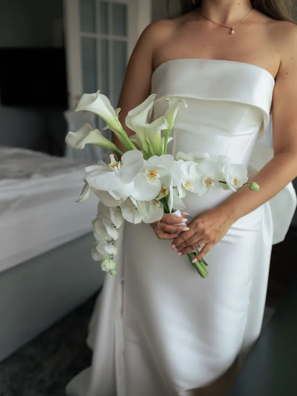 Braut in schlichtem weißem Kleid hält einen langen Strauß aus weißen Calla und Orchideen