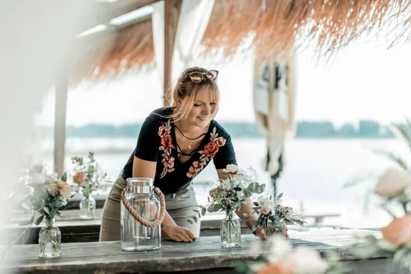 Floristin arrangiert kleine Gläser mit rosa und weißen Blumen auf einer Hochzeitstafel am Wasser
